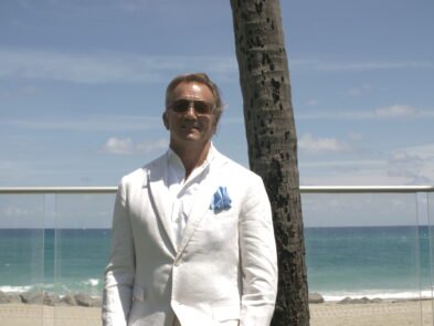 A man in white suit standing next to palm tree.